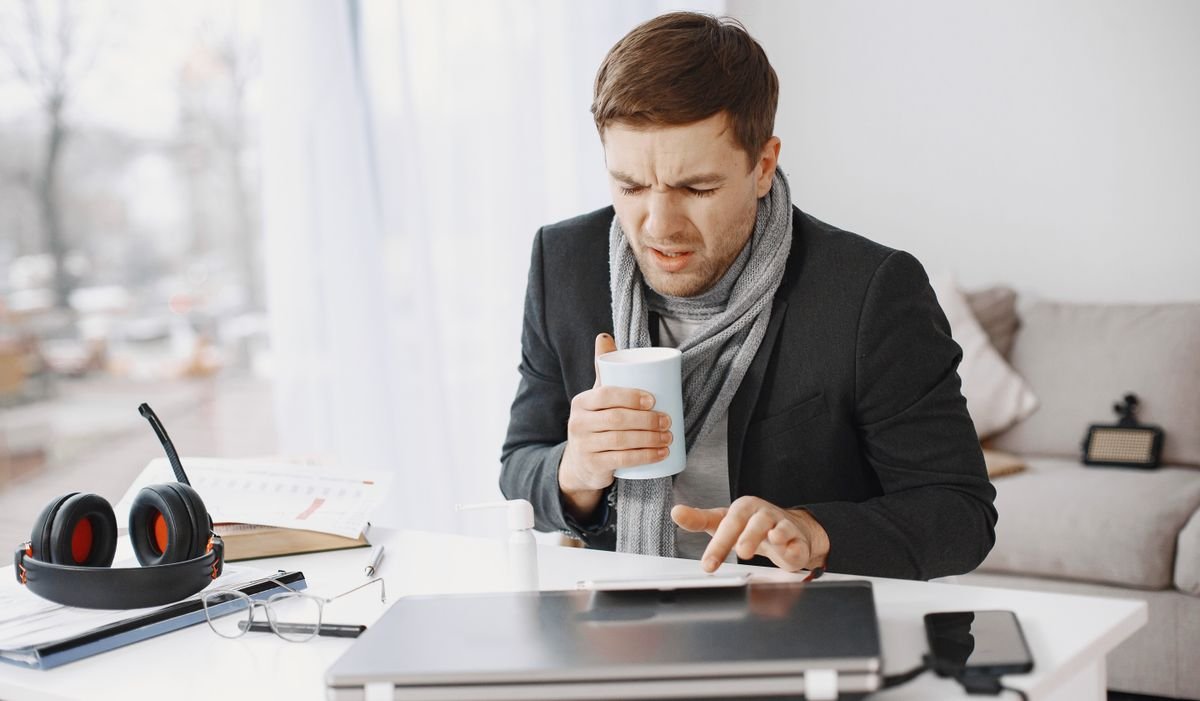 Trabajador enfermo con bufanda y una taza en su escritorio, preocupado por la incapacidad temporal y quién paga la baja